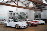 Inside a rustic garage with a high ceiling and white brick walls, three classic race cars are displayed on a wooden floor. Above the cars, spare parts and car accessories are arranged neatly against the wall. A framed painting of a car sits on an easel beside a potted plant.
