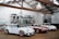 Inside a rustic garage with a high ceiling and white brick walls, three classic race cars are displayed on a wooden floor. Above the cars, spare parts and car accessories are arranged neatly against the wall. A framed painting of a car sits on an easel beside a potted plant.