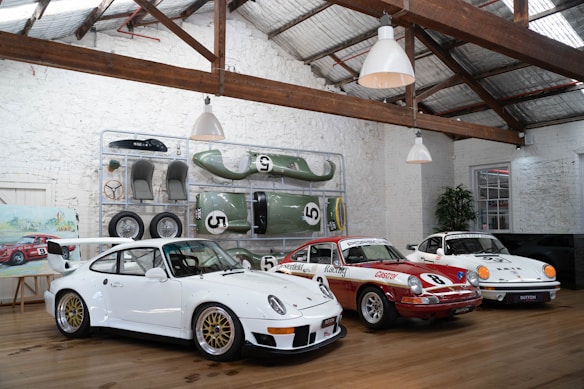 Inside a rustic garage with a high ceiling and white brick walls, three classic race cars are displayed on a wooden floor. Above the cars, spare parts and car accessories are arranged neatly against the wall. A framed painting of a car sits on an easel beside a potted plant.