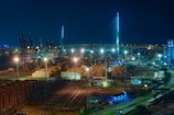A night-time view of a busy industrial port filled with stacks of shipping containers and illuminated by bright lights. Numerous cranes are visible throughout the port, and a large suspension bridge brightly lit contrasts against the dark sky. Buildings and structures are seen around the area, with roads leading into the port.