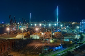 A night-time view of a busy industrial port filled with stacks of shipping containers and illuminated by bright lights. Numerous cranes are visible throughout the port, and a large suspension bridge brightly lit contrasts against the dark sky. Buildings and structures are seen around the area, with roads leading into the port.