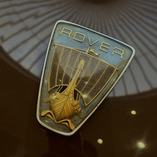 A vintage metallic emblem featuring a stylized Viking ship with a sail and intricate detailing. The word 'ROVER' is inscribed above the ship in a classic font. The emblem is set against a dark, glossy surface with reflections of surrounding architecture resembling a circular dome.