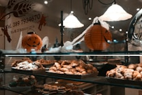 A bakery display case filled with a variety of pastries, including croissants and cookies. Above the pastries, there are Halloween-themed decorations, such as orange pumpkin lanterns with jack-o'-lantern faces and fake spider webs. The warm lighting creates a cozy atmosphere, and there is signage with contact information on the glass.