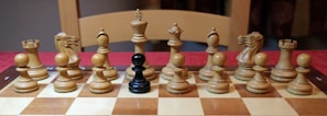 A chessboard with all wooden chess pieces arranged in the starting position, except for one black pawn placed in the center. The setting is indoors with a wooden chair visible in the background.
