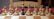 A chessboard with all wooden chess pieces arranged in the starting position, except for one black pawn placed in the center. The setting is indoors with a wooden chair visible in the background.