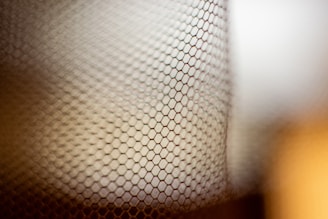 A close-up view of a mesh or net with hexagonal patterns, creating an abstract and textured appearance. The depth of field is shallow, with the background softly out of focus, blending warm and cool tones.