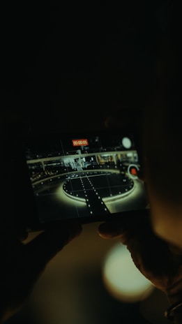 A person is holding a camera or a phone capturing an image of a circular stage or platform with lights around it, as seen through the viewfinder or screen. The area appears to be indoors with a dark ambiance.