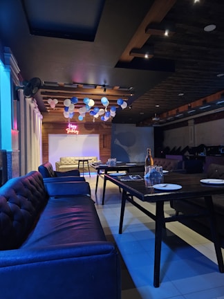 A dimly lit room with dark wooden ceiling panels and couches arranged around tables. Balloons in shades of blue, white, and light pink are suspended from the ceiling. A neon sign displays 'Happy Birthday'. Plates and glasses are set on the tables, suggesting a celebration setup.