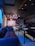 A dimly lit room with dark wooden ceiling panels and couches arranged around tables. Balloons in shades of blue, white, and light pink are suspended from the ceiling. A neon sign displays 'Happy Birthday'. Plates and glasses are set on the tables, suggesting a celebration setup.