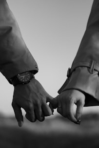 Two hands are intertwined, seemingly engaged in a gentle pinky promise. The right hand wears a watch, while the left hand is adorned with a ring. Both individuals are dressed in long-sleeved jackets.