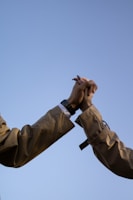 Two hands clasped together in a gesture of connection, set against a clear blue sky. The individuals are wearing light brown coats, and one hand has a watch.