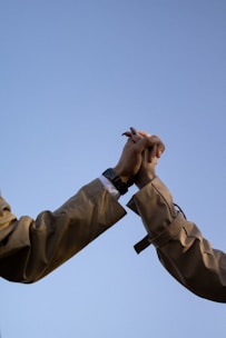 Two hands clasped together in a gesture of connection, set against a clear blue sky. The individuals are wearing light brown coats, and one hand has a watch.
