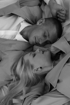 A black and white image of a man and a woman lying closely together, both wearing coats. They appear relaxed and content, with the woman resting her head on the man's shoulder. Their expressions suggest a sense of intimacy and tranquility.