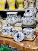 A collection of white ceramic containers adorned with intricate blue floral patterns is elegantly arranged. The set includes a vase, several lidded jars, and a tray, all placed on a wooden surface. In one jar, seeds are visible, while others are topped with lids featuring matching blue knobs. The background showcases more ceramic vases with various patterns and colors.