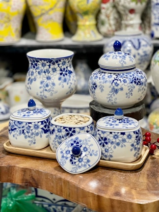 A collection of white ceramic containers adorned with intricate blue floral patterns is elegantly arranged. The set includes a vase, several lidded jars, and a tray, all placed on a wooden surface. In one jar, seeds are visible, while others are topped with lids featuring matching blue knobs. The background showcases more ceramic vases with various patterns and colors.
