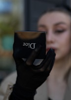 A person holds a compact makeup mirror from Dior in their hand. The hand is adorned with a black mesh glove. The person's face is partially visible and blurred in the background.