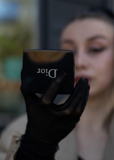 A person holds a compact makeup mirror from Dior in their hand. The hand is adorned with a black mesh glove. The person's face is partially visible and blurred in the background.