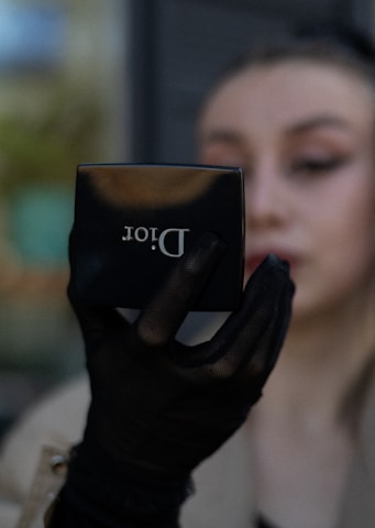 A person holds a compact makeup mirror from Dior in their hand. The hand is adorned with a black mesh glove. The person's face is partially visible and blurred in the background.