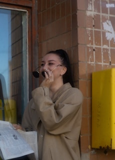 A person is standing outdoors against a brick wall. They are holding sunglasses in one hand and a newspaper in the other. The person is laughing, with their head slightly tilted back. A yellow box is mounted on the wall next to them, and a window is visible to the left.