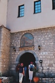 A stone building with an arched entrance featuring a mosaic above it, depicting religious figures. Several people wearing casual clothes and backpacks are seen entering the building. There are potted plants beside the entrance and the stone walls have lantern-style lights.
