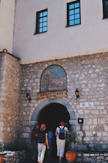 A stone building with an arched entrance featuring a mosaic above it, depicting religious figures. Several people wearing casual clothes and backpacks are seen entering the building. There are potted plants beside the entrance and the stone walls have lantern-style lights.