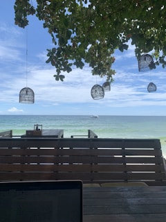 A serene beachfront view with clear blue skies and a calm ocean. Overhead, leafy branches with hanging basket lights create a shaded area. A wooden deck overlooks the sea, allowing for a relaxed setup with a laptop visible in the foreground.