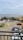 A parking area with several cars is situated near a coastal view. In the background, the ocean is visible with a ship on the horizon. Surrounding the parking lot are various tropical plants and a building structure.