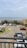 A parking area with several cars is situated near a coastal view. In the background, the ocean is visible with a ship on the horizon. Surrounding the parking lot are various tropical plants and a building structure.