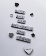 Wooden letter tiles arranged to form the question 'What are you grateful for today?' are scattered across a white surface. Decorative items such as a black heart, a grey heart, and a couple of shiny stones surround the message. One of the hearts has the handwritten name 'MarkC' on it.