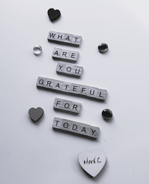 Wooden letter tiles arranged to form the question 'What are you grateful for today?' are scattered across a white surface. Decorative items such as a black heart, a grey heart, and a couple of shiny stones surround the message. One of the hearts has the handwritten name 'MarkC' on it.