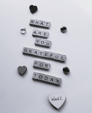 Wooden letter tiles arranged to form the question 'What are you grateful for today?' are scattered across a white surface. Decorative items such as a black heart, a grey heart, and a couple of shiny stones surround the message. One of the hearts has the handwritten name 'MarkC' on it.