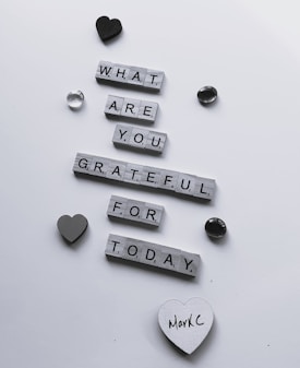 Wooden letter tiles arranged to form the question 'What are you grateful for today?' are scattered across a white surface. Decorative items such as a black heart, a grey heart, and a couple of shiny stones surround the message. One of the hearts has the handwritten name 'MarkC' on it.
