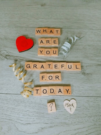 Wooden Scrabble tiles spell out the phrase 'What are you grateful for today?' on a light wooden surface. Two decorative curling ribbons, one gold and one silver, are positioned around the text. A small red heart and a white heart-shaped paper with written text are included in the scene.