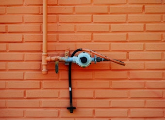 An orange brick wall with a metal gas pipe attached to it. The pipe is connected to a valve with a pressure gauge, and a hose extending horizontally. The wall features a repeating rectangular brick pattern.