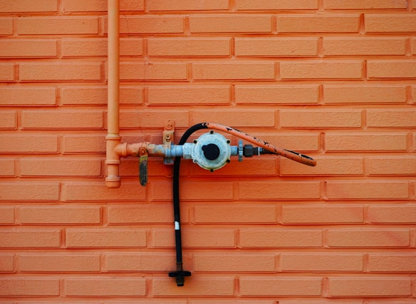 An orange brick wall with a metal gas pipe attached to it. The pipe is connected to a valve with a pressure gauge, and a hose extending horizontally. The wall features a repeating rectangular brick pattern.