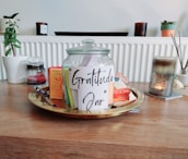 A glass jar labeled 'Gratitude Jar' is placed on a wooden table, surrounded by various items including books, a pen, and a candle. The jar is placed on a round tray. In the background, there are potted plants and additional candles on a shelf.