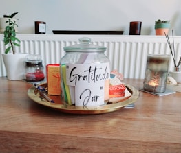A glass jar labeled 'Gratitude Jar' is placed on a wooden table, surrounded by various items including books, a pen, and a candle. The jar is placed on a round tray. In the background, there are potted plants and additional candles on a shelf.