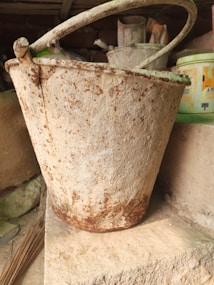 An old, rusty metal bucket with a handle is placed on a concrete slab. The bucket has visible signs of wear, with rust patches and a weathered appearance. In the background, there are other containers with faded labels and signs of age.