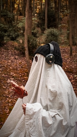 A person dressed as a ghost with a white sheet draped over them stands in a forest. They are wearing a black beanie hat and headphones, holding a dried autumn leaf. Dark glasses are visible through holes in the sheet. The forest floor is covered in fallen leaves, and tall trees surround the scene.