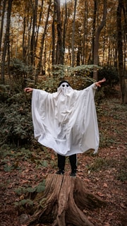 A person dressed as a ghost with a white sheet covering them, standing with arms outstretched on a tree stump. They are wearing black sunglasses, surrounded by a dense forest with tall trees and a scattering of fallen leaves.