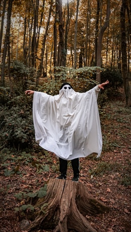 A person dressed as a ghost with a white sheet covering them, standing with arms outstretched on a tree stump. They are wearing black sunglasses, surrounded by a dense forest with tall trees and a scattering of fallen leaves.
