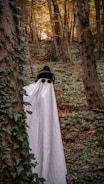 A person is dressed in a makeshift ghost costume, featuring a white sheet with eye cutouts, standing among trees in a densely wooded forest filled with green foliage. They are also wearing a black knit hat and headphones, adding a playful contrast to the spooky theme. Ivy climbs along the tree trunks, and the autumn leaves provide a backdrop of warm hues.