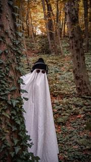 A person is dressed in a makeshift ghost costume, featuring a white sheet with eye cutouts, standing among trees in a densely wooded forest filled with green foliage. They are also wearing a black knit hat and headphones, adding a playful contrast to the spooky theme. Ivy climbs along the tree trunks, and the autumn leaves provide a backdrop of warm hues.