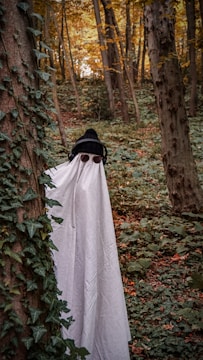 A person is dressed in a makeshift ghost costume, featuring a white sheet with eye cutouts, standing among trees in a densely wooded forest filled with green foliage. They are also wearing a black knit hat and headphones, adding a playful contrast to the spooky theme. Ivy climbs along the tree trunks, and the autumn leaves provide a backdrop of warm hues.