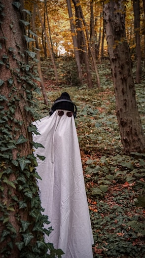 A person is dressed in a makeshift ghost costume, featuring a white sheet with eye cutouts, standing among trees in a densely wooded forest filled with green foliage. They are also wearing a black knit hat and headphones, adding a playful contrast to the spooky theme. Ivy climbs along the tree trunks, and the autumn leaves provide a backdrop of warm hues.