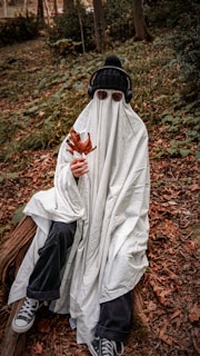 A person dressed as a ghost sitting on a log in a forest setting. The costume consists of a white sheet covering the entire body, with sunglasses and headphones worn over the sheet. The person holds a large dry leaf, and the ground is covered with fallen leaves, surrounded by green shrubbery and trees.