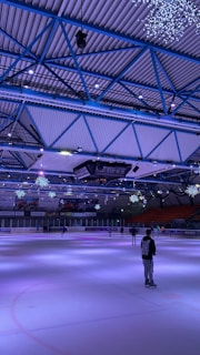 An indoor ice skating rink with a high ceiling supported by blue steel beams. Snowflake-like decorations hang from the ceiling, and the arena is dimly lit, creating a calming atmosphere. Several people are skating on the ice, with one person in the foreground wearing a black hoodie with a white design on the back.