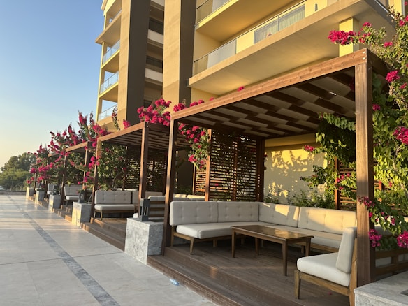 A modern outdoor seating area with wooden pergolas adorned with vibrant pink flowers. The seating consists of white cushioned benches arranged around wooden tables. The background features a contemporary building with large balconies, basking in the warm glow of the late afternoon sun.