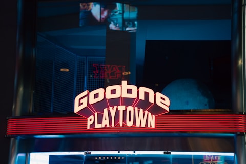 A neon sign reading 'Goobne Playtown' is prominently displayed against a dark background. The sign is illuminated with a bright pinkish-red color and features an art deco style with horizontal lines crossing beneath the text. In the background, there are reflections and faint images on glass, suggesting an indoor setting with dim lighting.