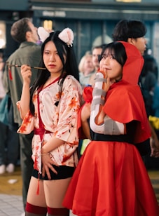 Two women are dressed in elaborate costumes, suggesting a cosplay or festival event. One woman wears a floral-patterned kimono with cat ears and holds a decorative pipe. The other is in a red cloak with a hood, resembling a storybook character. Both expressions vary, with one looking serious and the other appearing contemplative. The background is a bustling environment, likely a street or convention, with other people blurred in the background.